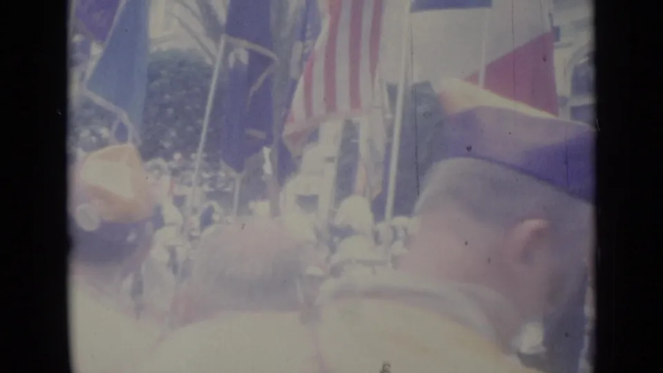 1962 Nice parade scene with spectators in the foreground, several flags overhead, and uniformed marchers visible through the crowd.