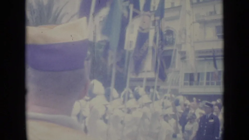 Nice, France, 1962 frame showing a handheld crowd view of uniformed marchers, flags, and nearby spectators during a street parade.