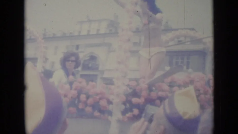 A woman rides on the flower-covered float in Nice, with spectators and a pale urban facade visible in the background.