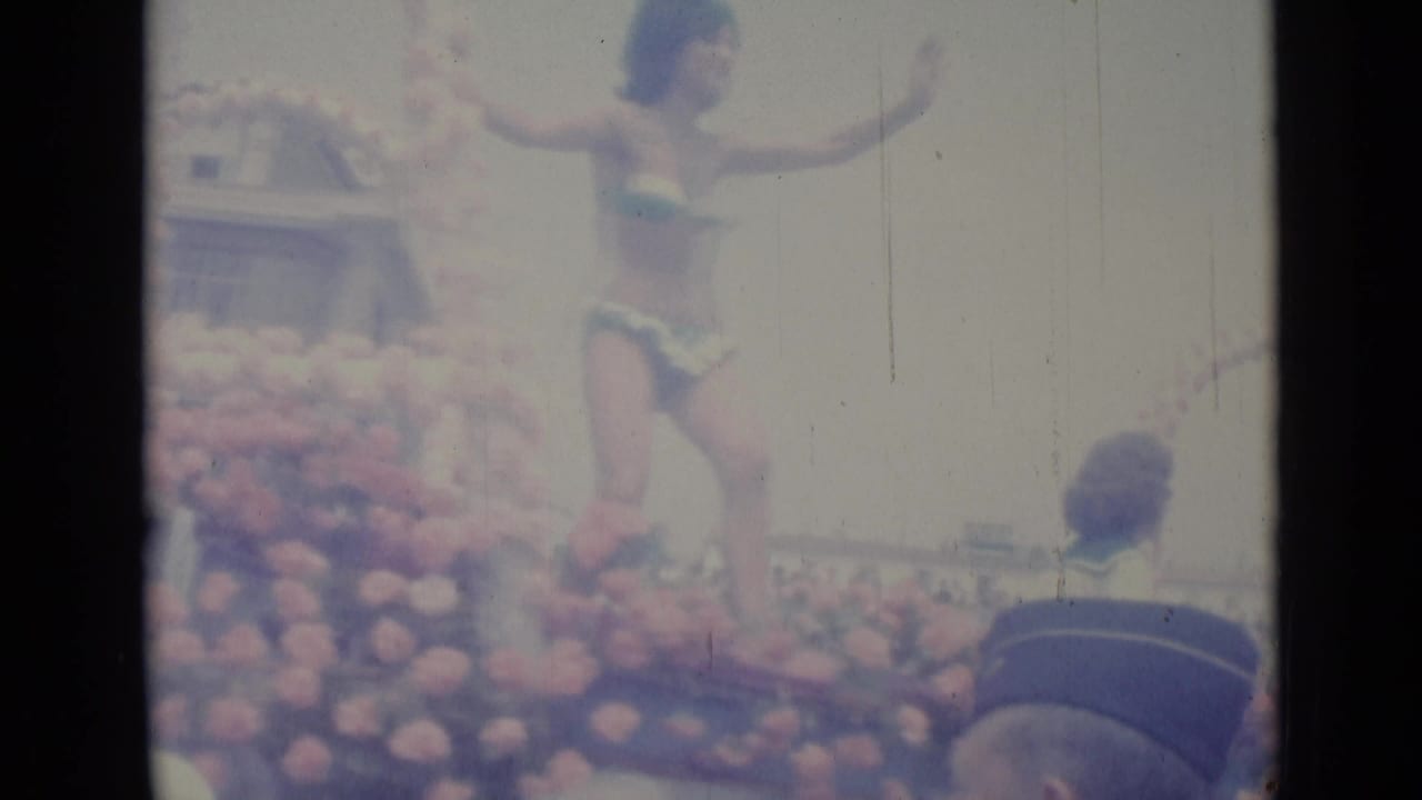 Woman in a bikini waves from a flower-covered parade float before a crowded street.