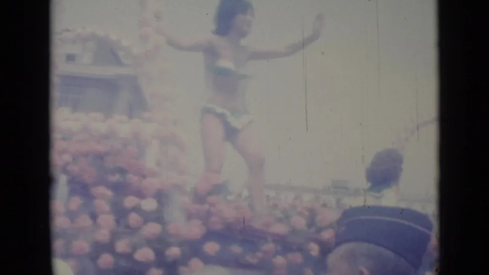 1962 Nice parade frame showing a bikini-clad woman waving on a floral float with spectators packed along the route.