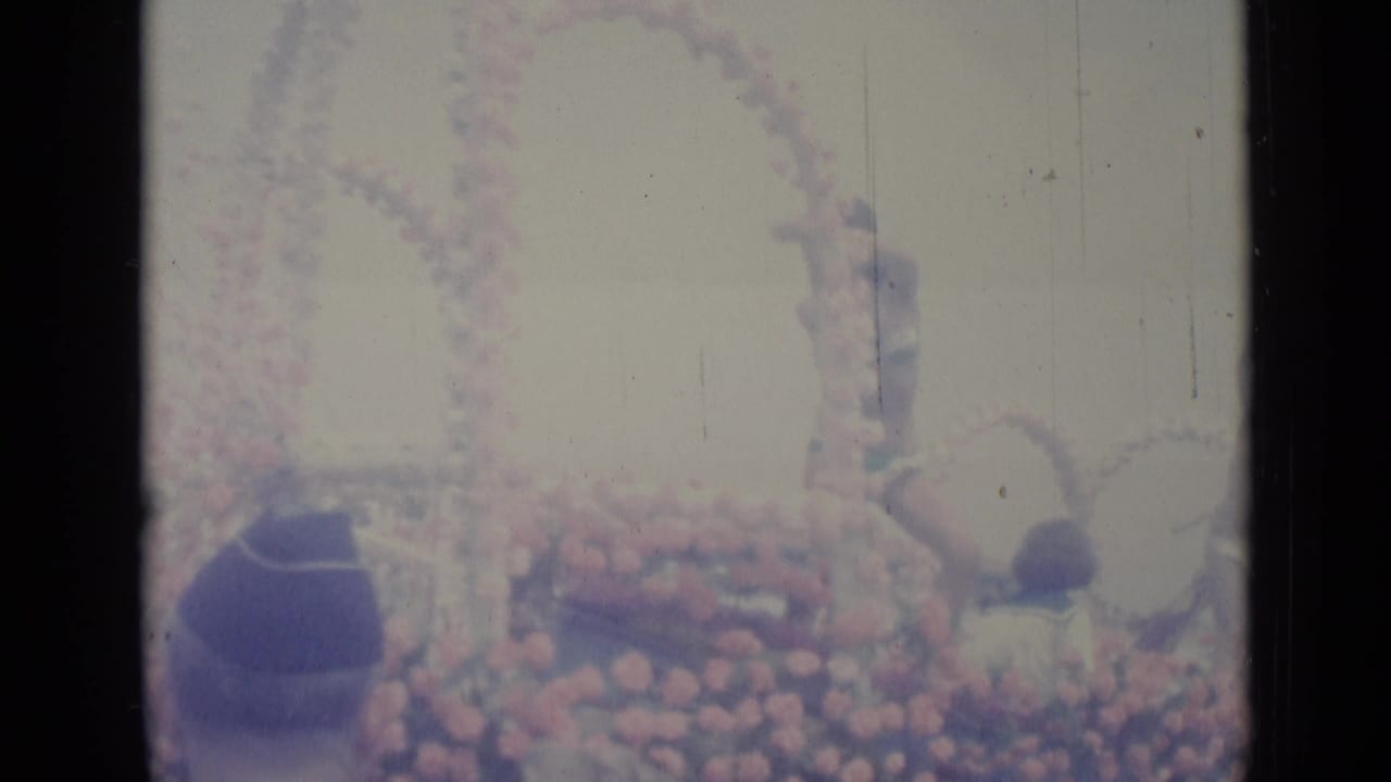 Floral parade float passes a crowded street with a seated participant in the foreground.