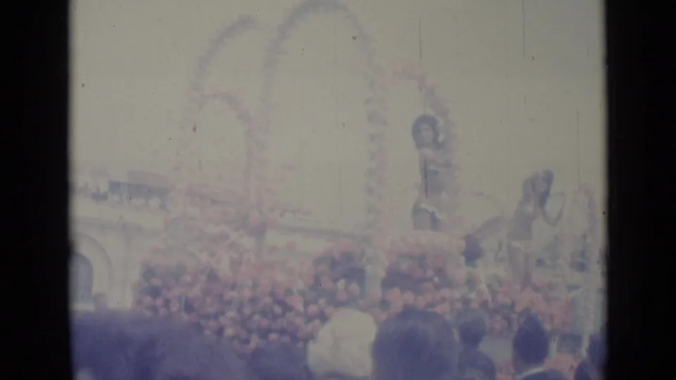 1962 Nice parade frame with a flower-covered float, a standing woman, another rider, and a crowd seen through faded 8mm exposure.