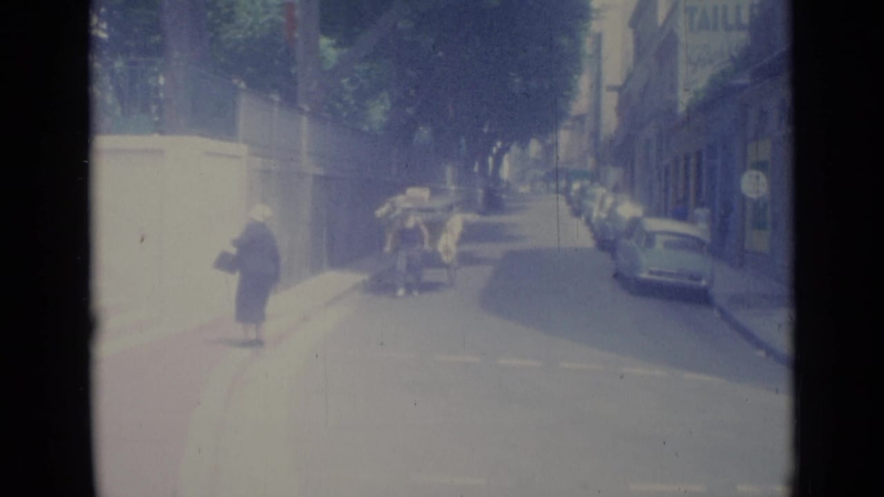 Sunlit street with a woman on the sidewalk, parked cars, and a loaded cart in the road.