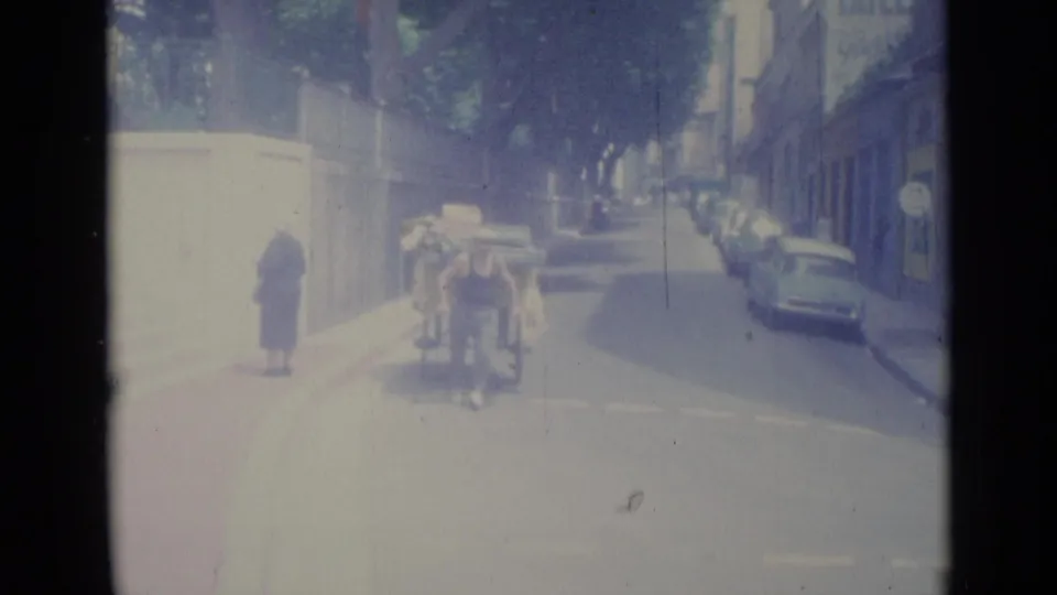 A man appears to push a loaded cart along a sunlit Nice street, with a woman and parked cars in view.