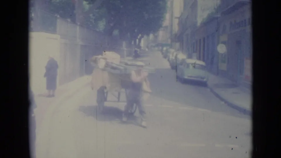 A man in a sleeveless shirt pushes a tall loaded cart across a 1962 Nice street with cars parked behind him.