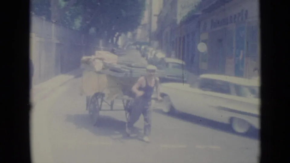 In Nice, France, a man moves a heavily loaded hand cart along a sunlit city street in 1962, with mid-century cars nearby.