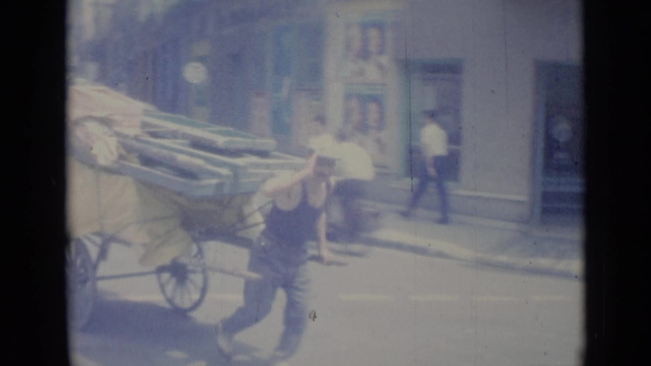 Man walking beside a loaded cart as a light-colored car passes on the street.