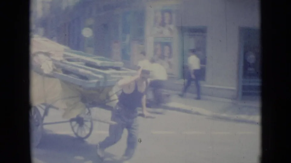 The cart pusher continues along a Nice street in 1962 while a pale mid-century car and pedestrians appear in the background.