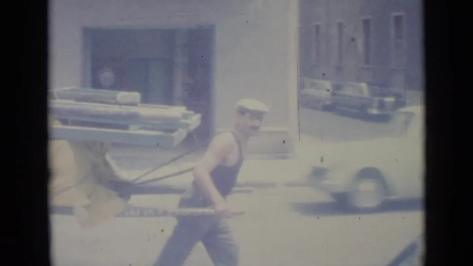A closer 8mm view shows the man leaning into the cart handles on a bright Nice street, with a moving car blurred in the background.