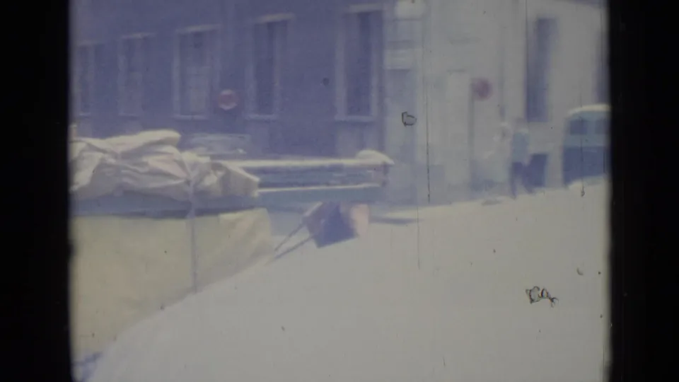 The loaded hand cart passes toward the edge of the frame on a Nice street, with building fronts and distant pedestrians visible.