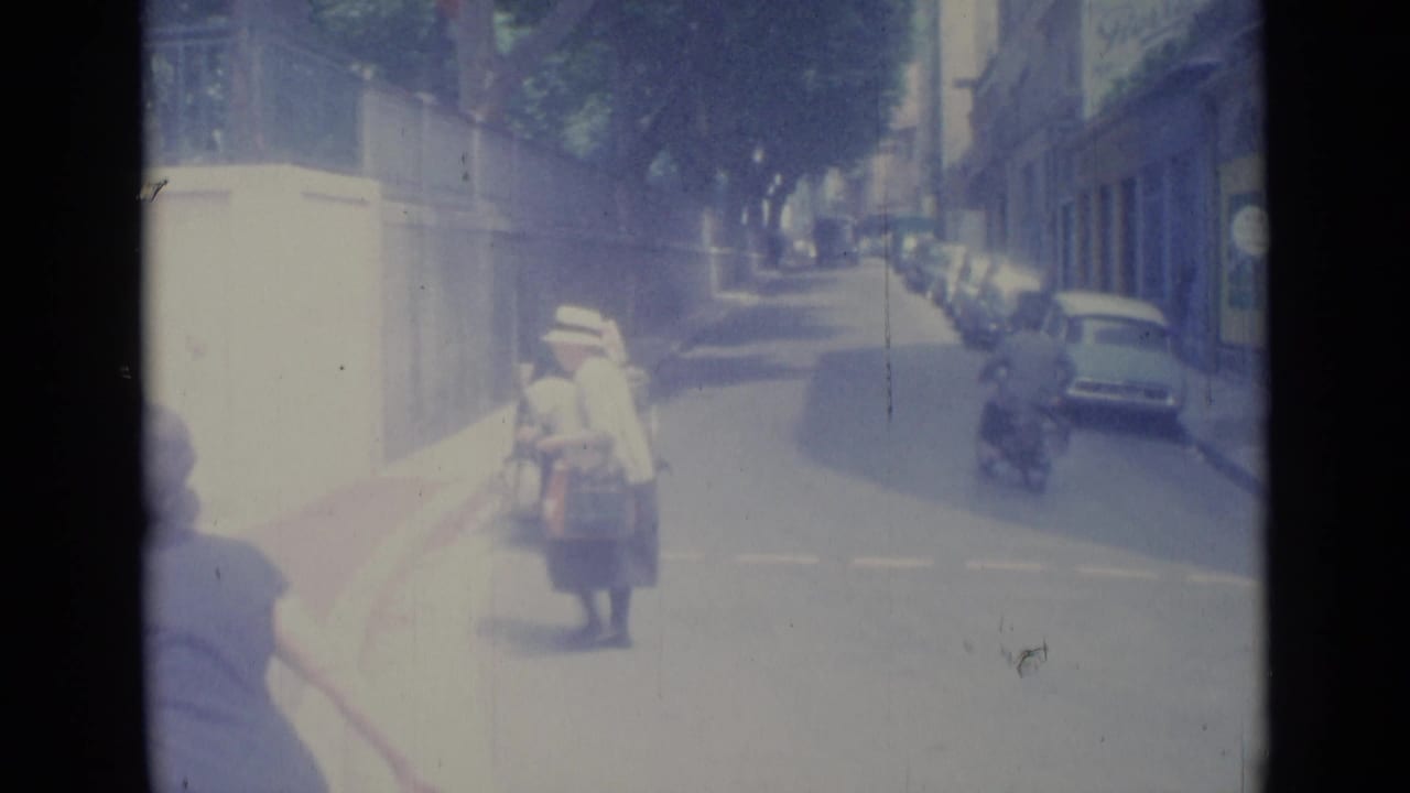 Older woman and another pedestrian near a sunlit street with parked cars and a motor scooter.