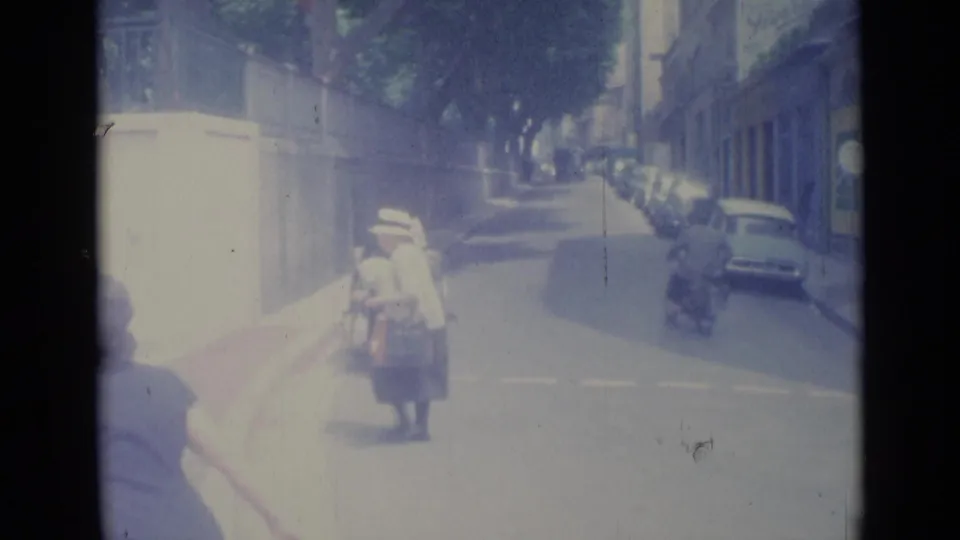 1962 Nice street view with pedestrians near the curb, parked cars along the right side, and a motor scooter farther down the road.