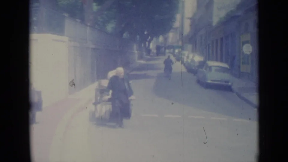 In Nice, France, an older woman in dark clothing moves beside a loaded handcart on a bright street with parked cars and a scooter.