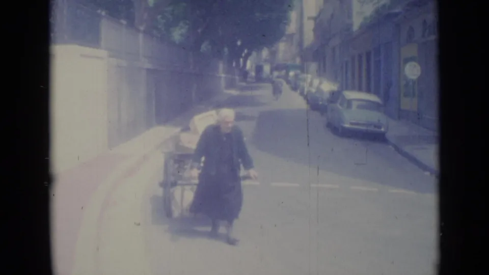1962 Nice footage shows an older woman in dark clothing walking near a loaded handcart, with parked cars and shaded buildings behind her.