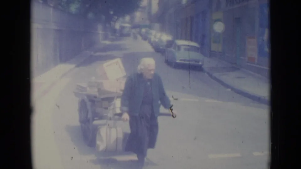 An older woman in dark clothing passes close to the camera on a Nice street, with a loaded handcart, parked cars, and road markings behind her.