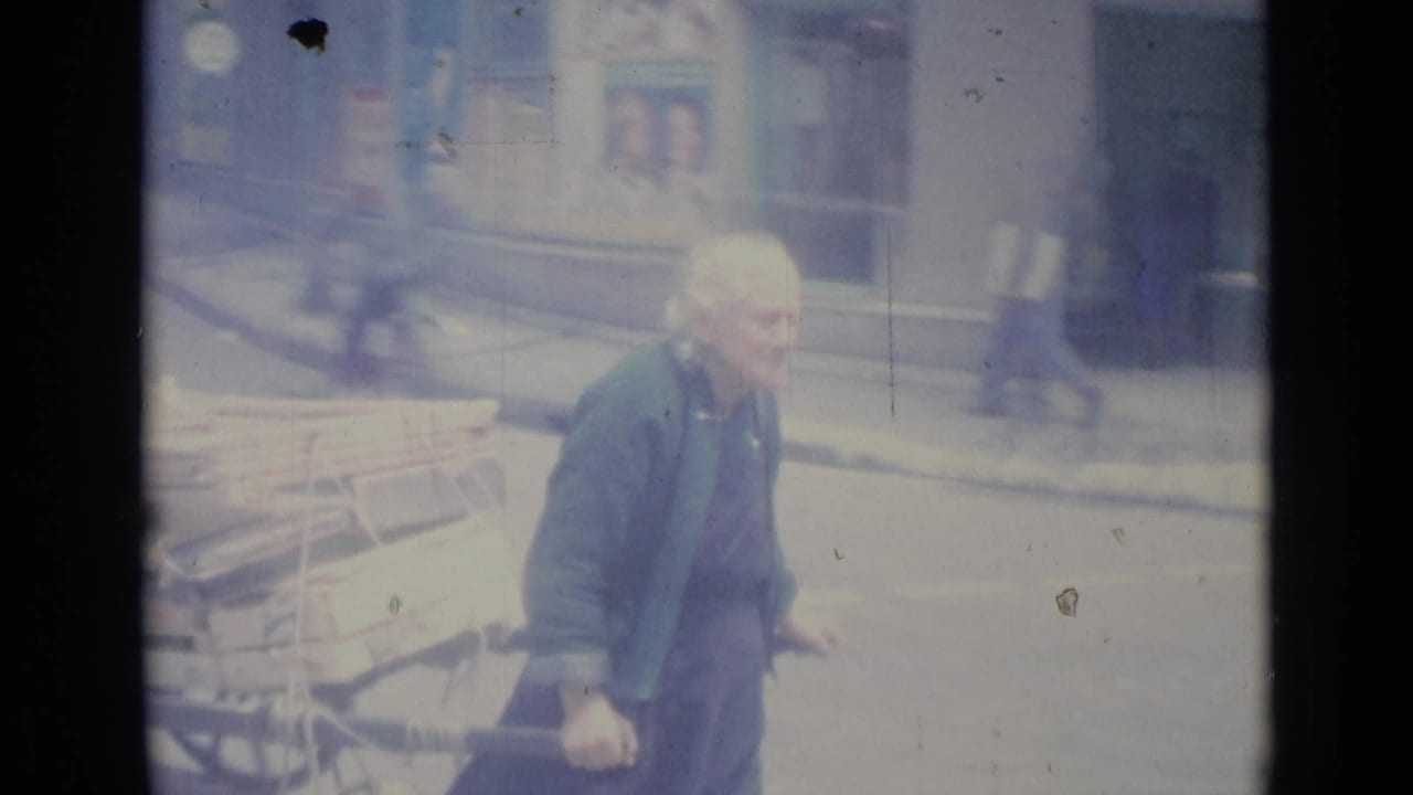 Older man beside a loaded handcart with pedestrians and buildings behind him.