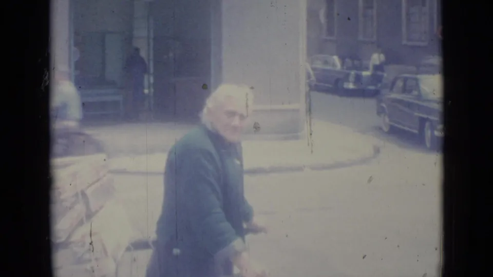 Older man with a loaded cart passes a doorway and parked cars in a 1962 Nice street view.