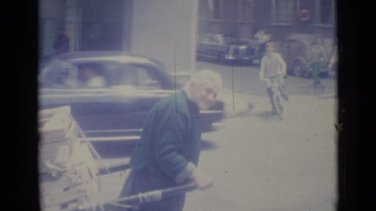 Elderly man pushes a loaded cart along a sunny street with cars behind him.