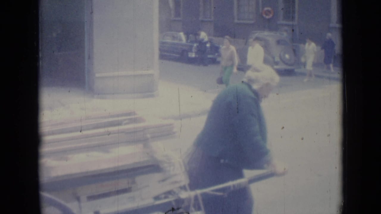The man continues pushing the cart beside a street with pedestrians and parked cars.