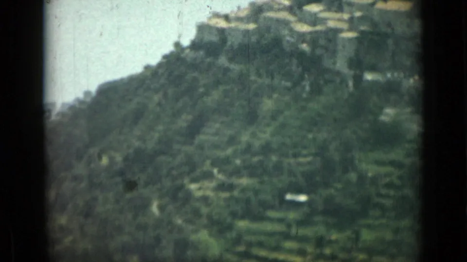 1962 Nice, France hillside scene with terraced green slopes, dense vegetation, and pale buildings seen through soft, faded 8mm color.