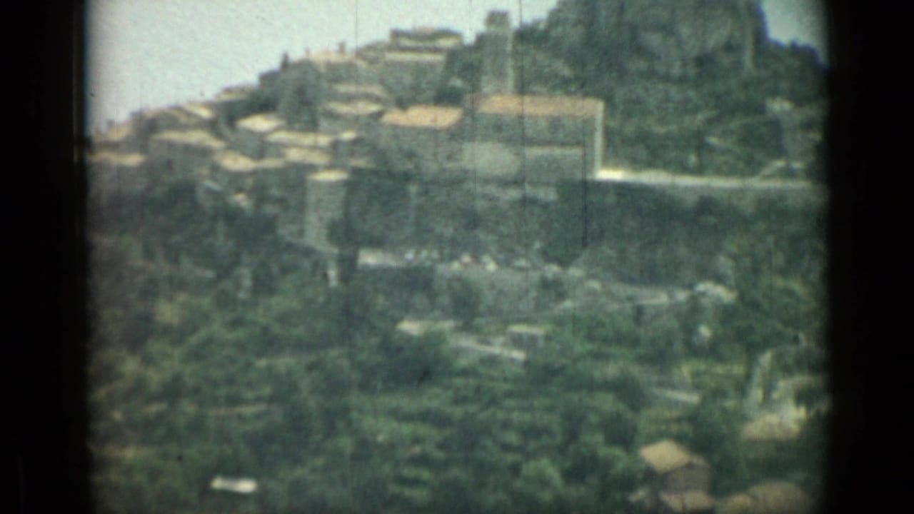 1962 Nice, France home-movie frame showing a hillside settlement, winding pale road, green groves, and rugged terrain.