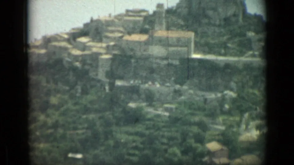 1962 Nice, France home-movie frame showing a hillside settlement, winding pale road, green groves, and rugged terrain.
