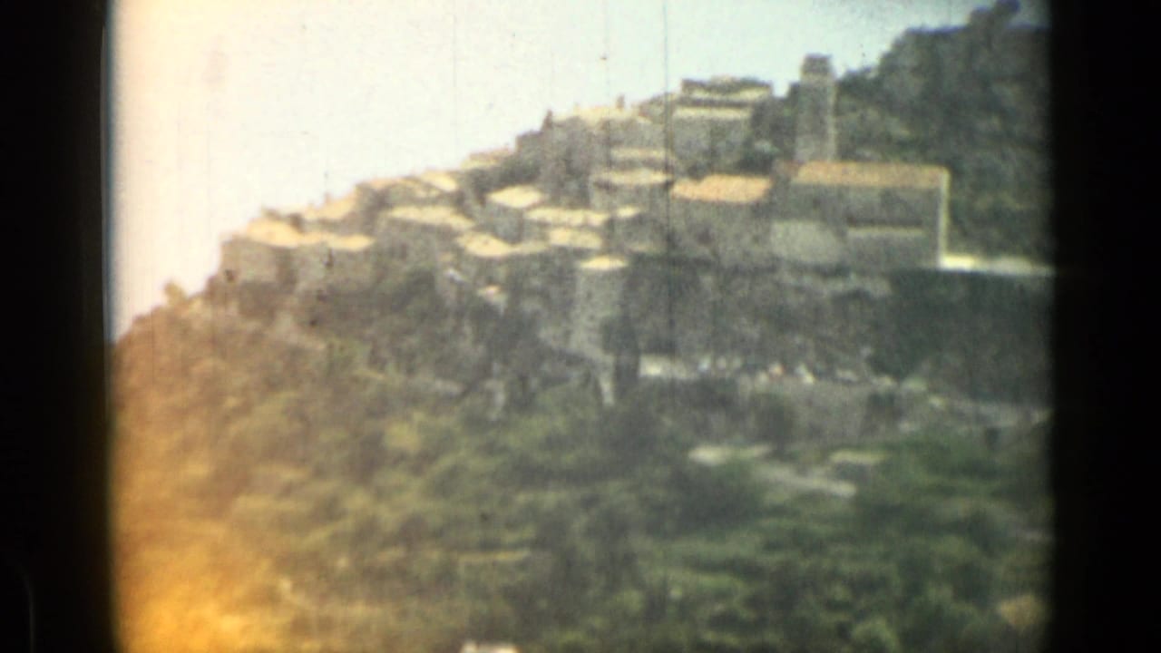 1962 8mm view near Nice, France, with sunlit dry brush in the foreground and a compact hillside village beyond.