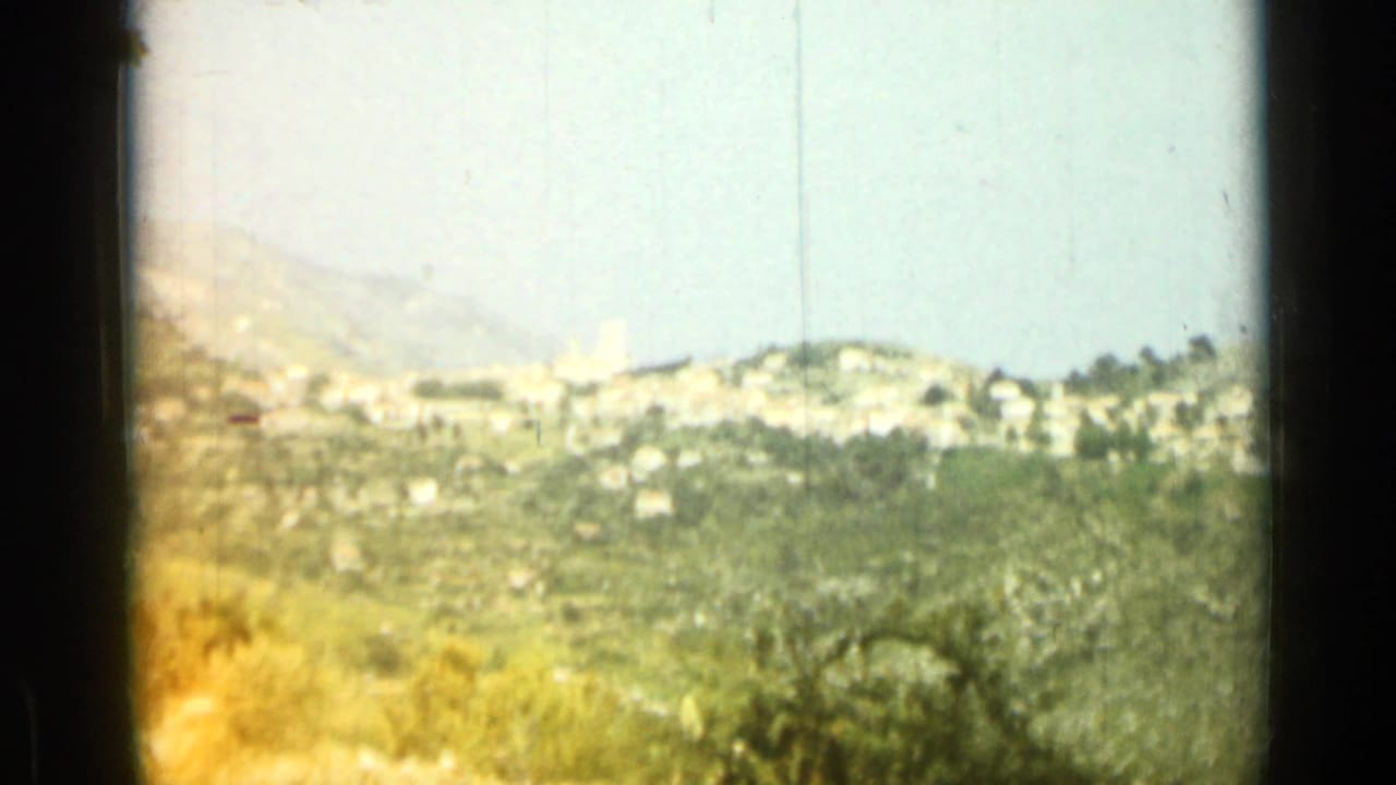 1962 Nice, France home-movie frame showing a wider hillside view with distant buildings, groves, dry brush, and hazy mountains.