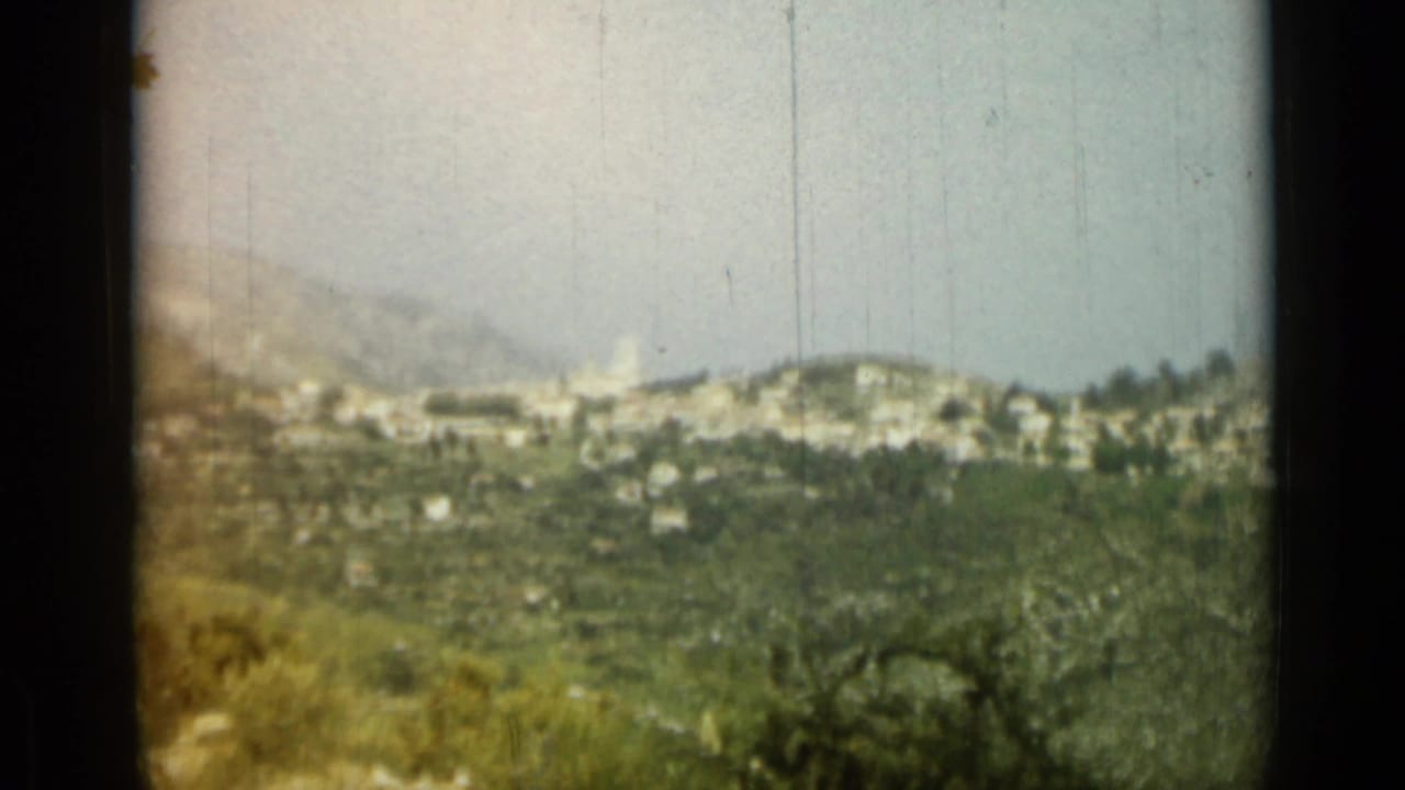 Distant hillside town with green slopes and pale buildings under a hazy sky.
