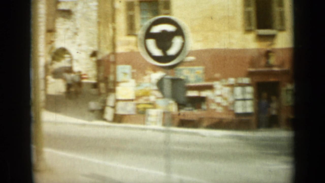 Street corner with a round traffic sign, shopfront displays, and stone buildings.