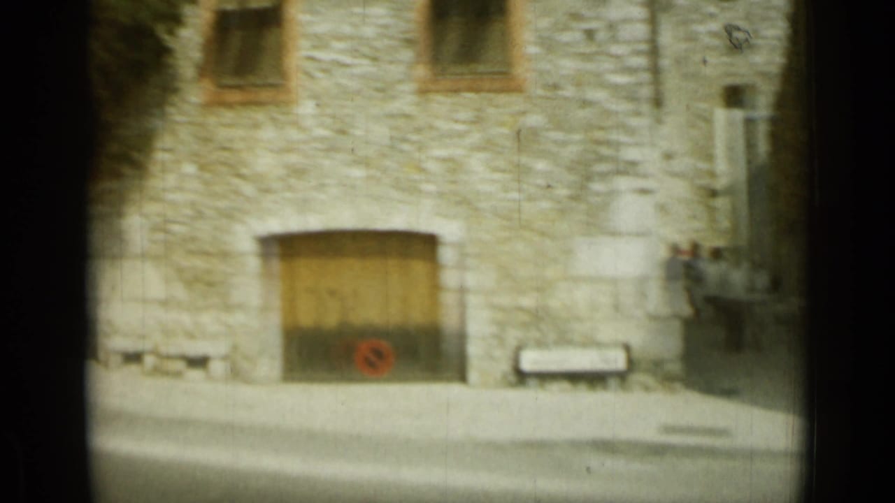 Stone facade in Nice with a broad wooden doorway, upper windows, a bench, and small roadside details in 1962 8mm footage.