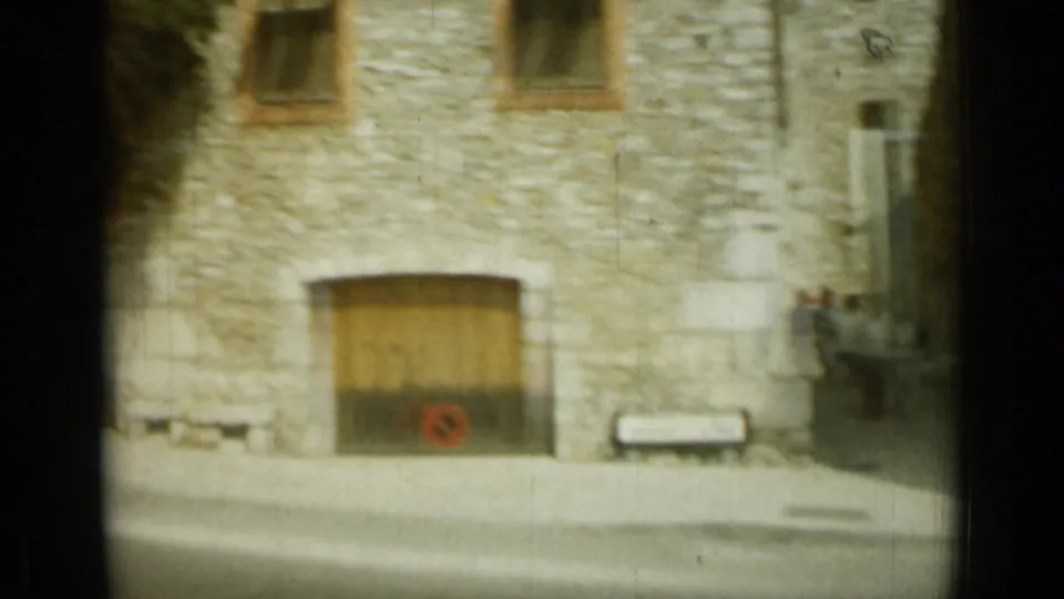Stone facade in Nice with a broad wooden doorway, upper windows, a bench, and small roadside details in 1962 8mm footage.