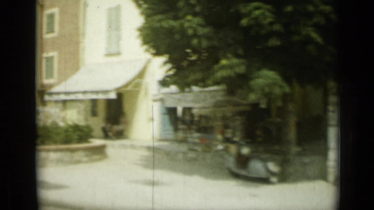 A shaded Nice plaza or side street in 1962 shows awnings, trees, stone walls, and parked vehicles in soft 8mm color.