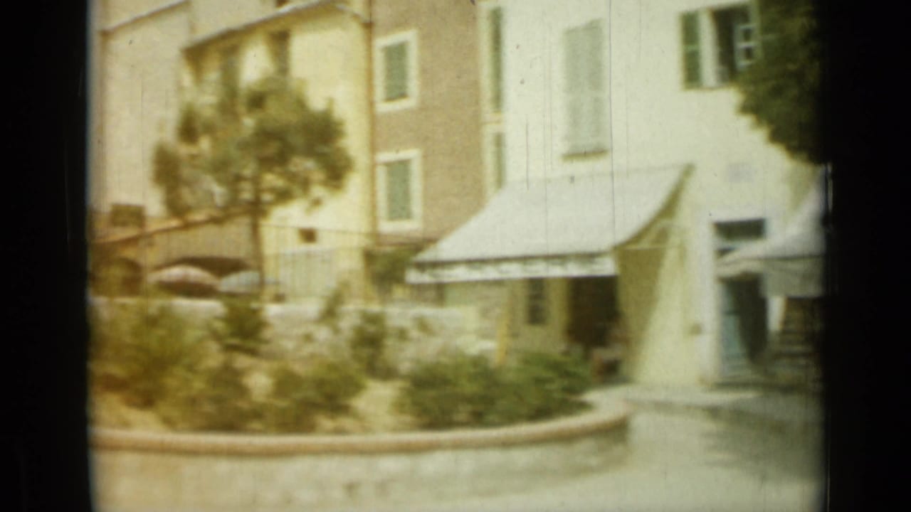 Soft street view with pale buildings, a tree, shrubs, and a storefront awning.