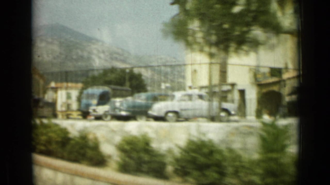Nice street scene with mid-century parked cars, a blue bus or van, pale buildings, trees, and distant hills.