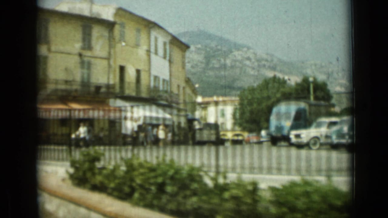 1962 Nice street corner seen through soft 8mm motion, with pedestrians, parked vehicles, railings, awnings, and hills behind the town.
