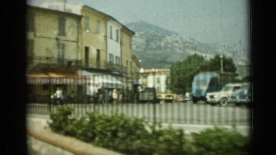 1962 Nice street corner seen through soft 8mm motion, with pedestrians, parked vehicles, railings, awnings, and hills behind the town.