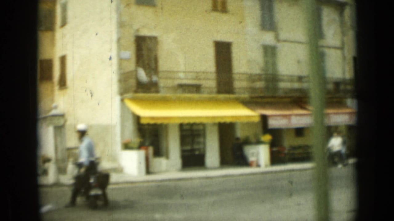 Street corner with pale buildings, a yellow awning, and a scooter rider passing at left.