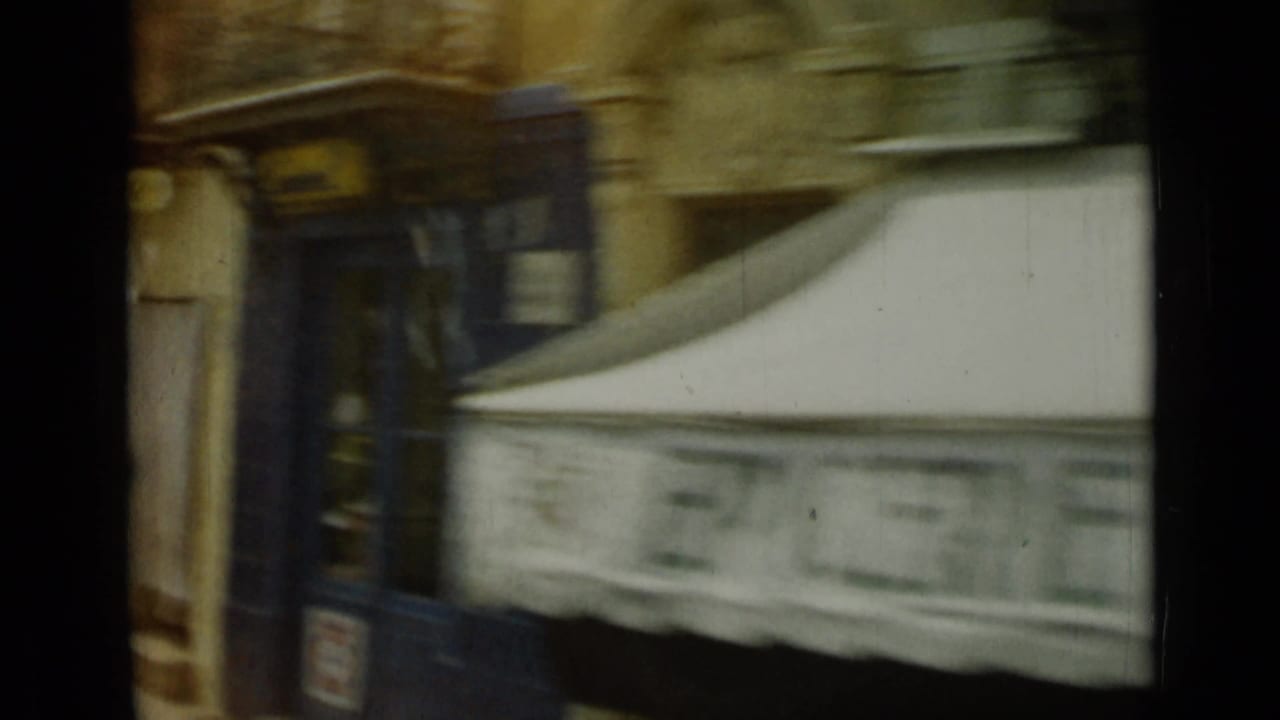 1962 Nice street detail with a blue storefront or kiosk, pale awning, and blurred masonry facade from a moving vehicle.