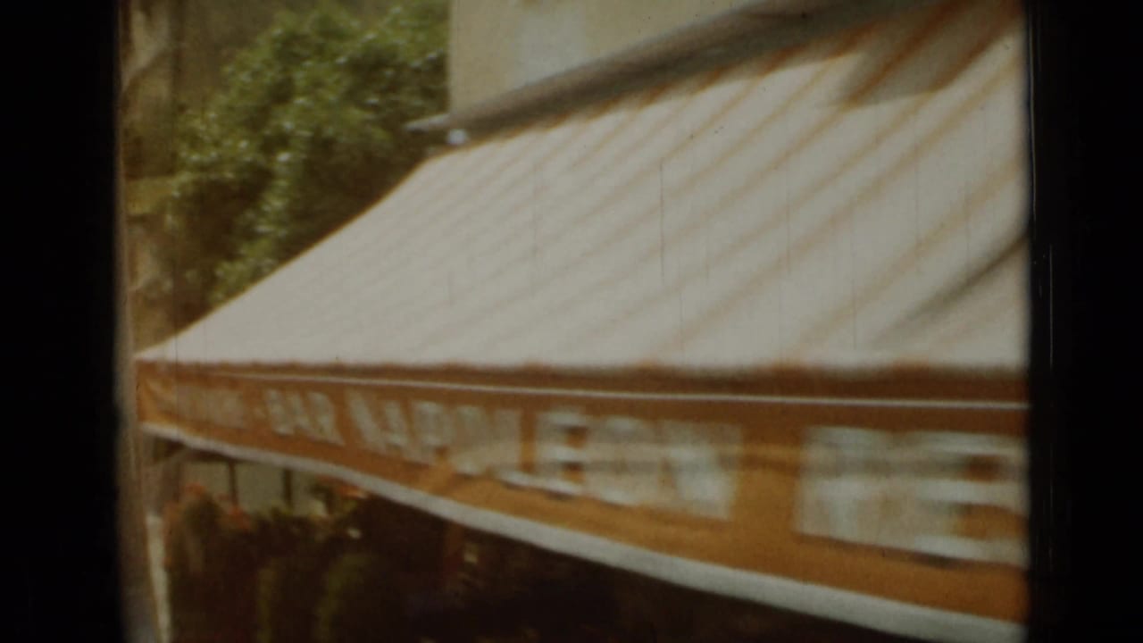 1962 Nice street-side awning seen from a moving camera, with orange-and-white stripes and surrounding foliage.