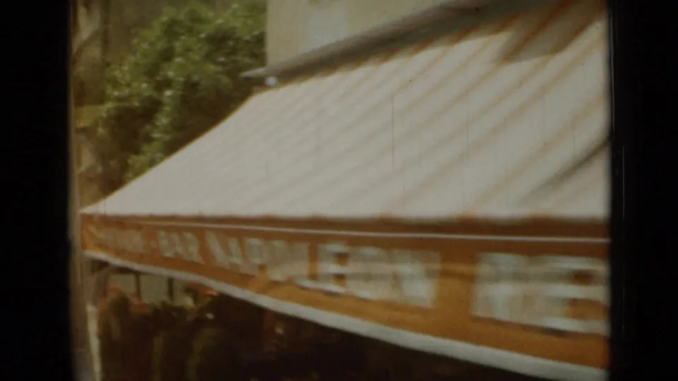 1962 Nice street-side awning seen from a moving camera, with orange-and-white stripes and surrounding foliage.