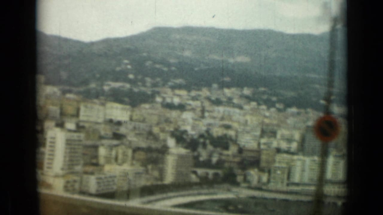 Elevated moving-vehicle view across Nice, France, with hillside buildings, a road barrier, and a small foreground sign visible.