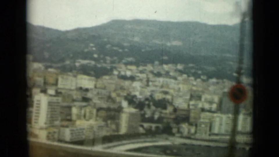 Elevated moving-vehicle view across Nice, France, with hillside buildings, a road barrier, and a small foreground sign visible.