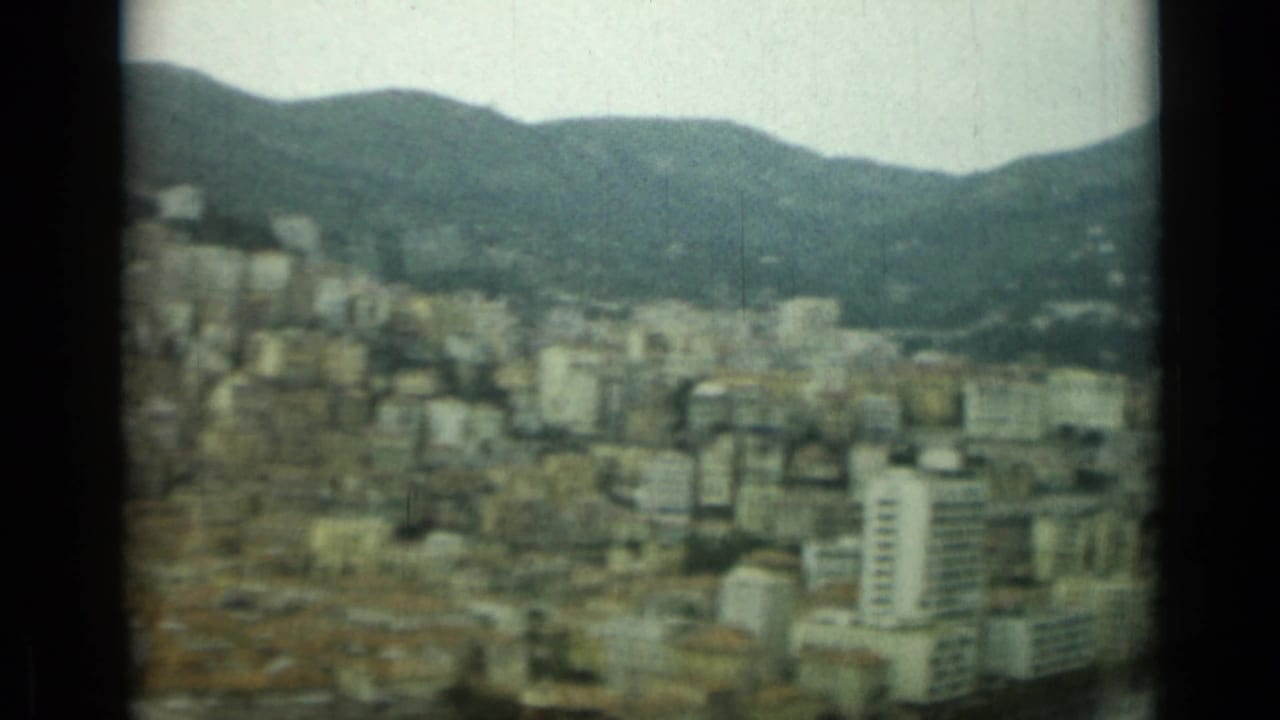 1962 home-movie frame showing Nice, France, spread across a hillside, with apartment blocks and mountains in soft focus.