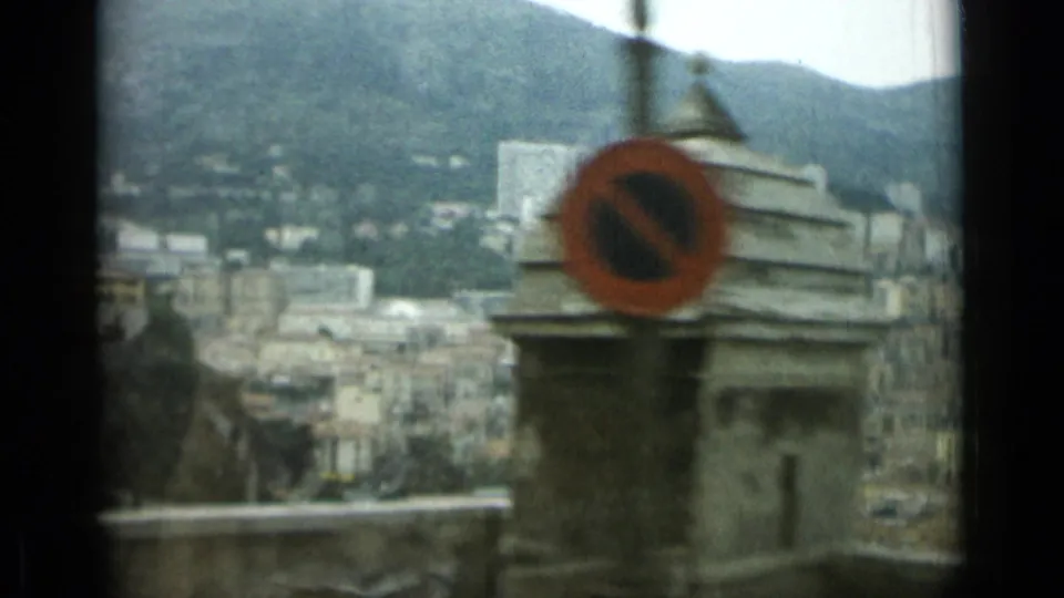1962 8mm view over Nice, France, with mountains behind city buildings and a roadside no-parking sign in the foreground.