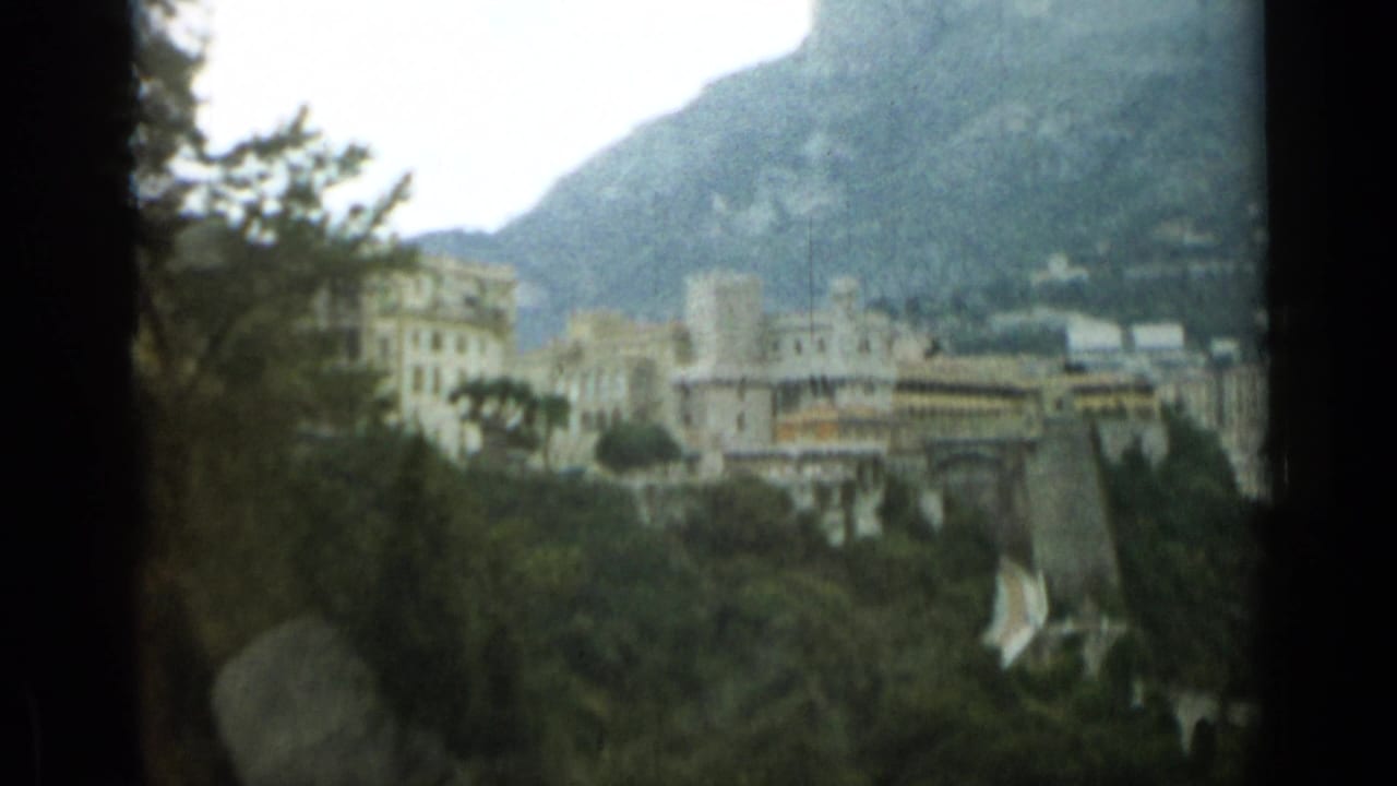 Soft 8mm frame of Nice in 1962, with foreground trees, pale buildings, and a mountain backdrop.