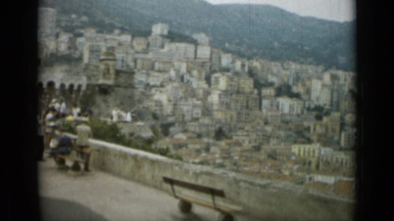 Tourists appear near wooden benches on a Nice overlook, with hillside buildings and mountains beyond.