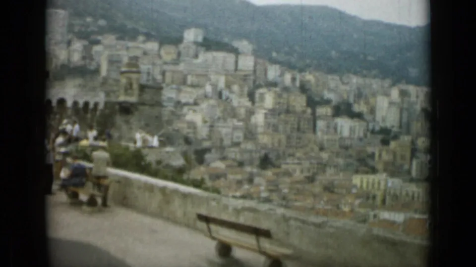Tourists appear near wooden benches on a Nice overlook, with hillside buildings and mountains beyond.