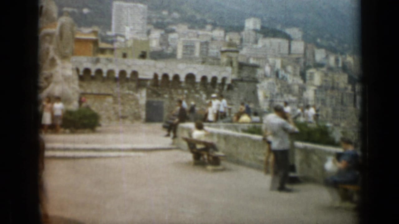 Tourists sit and stand near a stone monument, benches, and a crenellated wall above a cityscape.